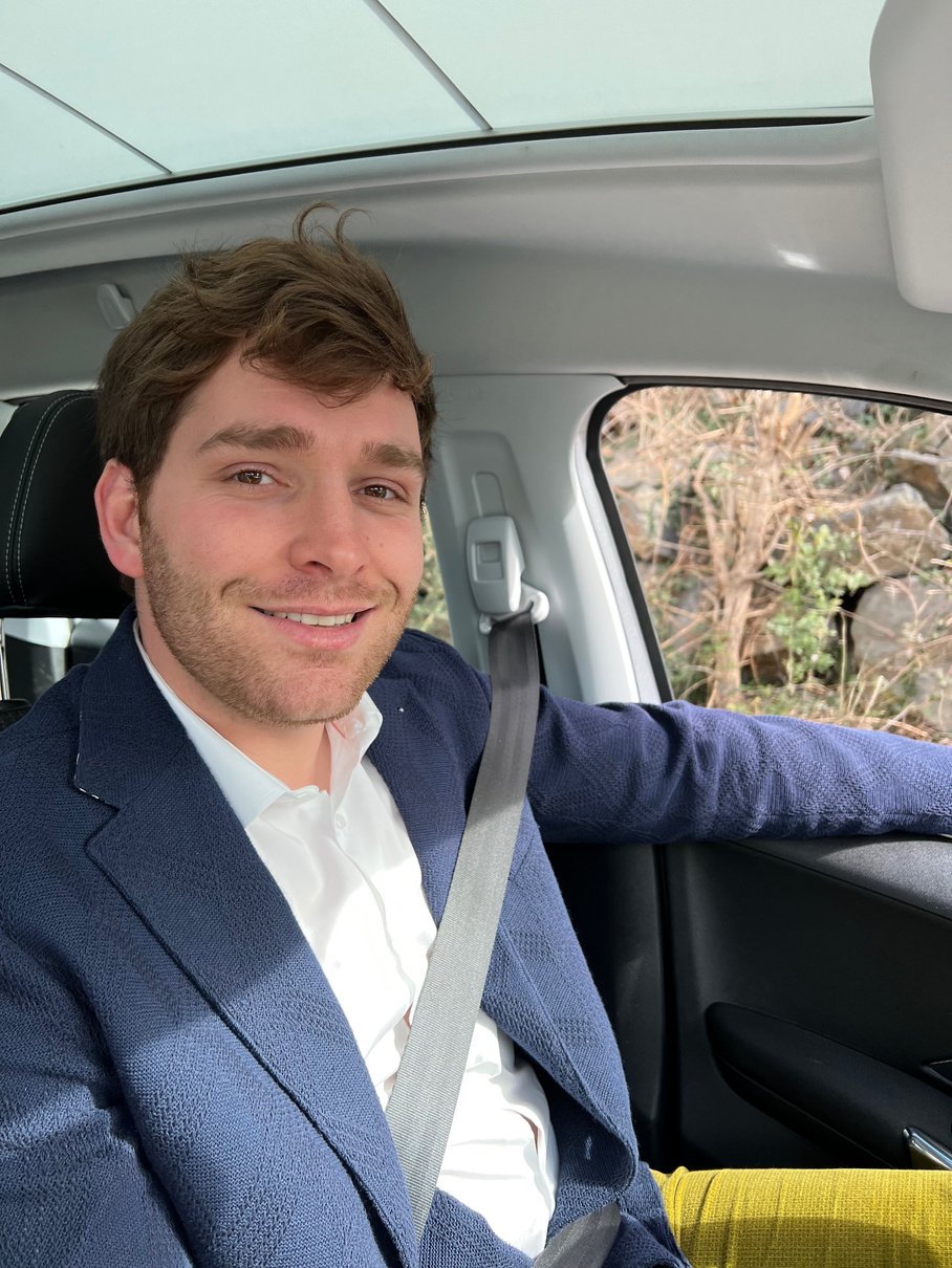 Photo de Jacques, votre agent automobile. Il est assis dans sa voiture avec la ceinture de sécurité attachée. Jacques vous sourit, il a mis son plus beau costume pour se présenter à vous.